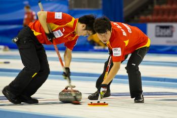 Two teammates bent over with their heads together, sweeping in front of a rock as it slides down the ice