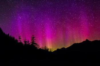 The night sky almost entirely lit up in neon pink and purple streaks over a silhouette of mountains and trees below