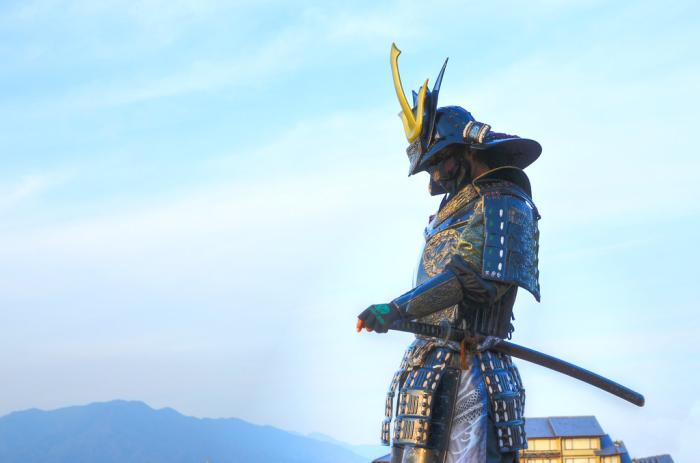 A man in samurai armor stands with his hand on his sword in front of a blue sky